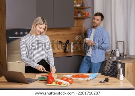 Beautiful young couple is talking and smiling while cooking healthy food in kitchen at home. Man is drinking cup of coffee and talking with woman