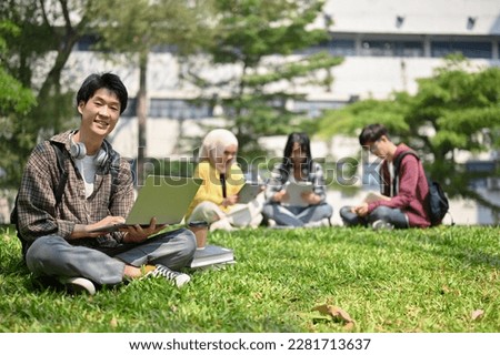 Smiling and happy young Asian male college student in flannel shirt sits on the grass in the campus park with his portable laptop. Royalty-Free Stock Photo #2281713637