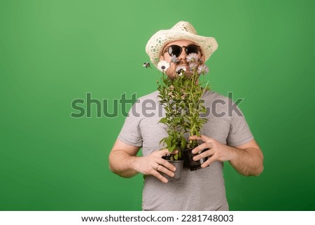 A male gardener, florist in a hat and sunglasses holds fresh flowers in pots on a green background. Plant breeding, sale.