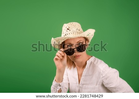 Cheerful amazed woman in a hat and sunglasses in a white shirt on a green background. Looks into the camera with his glasses down.