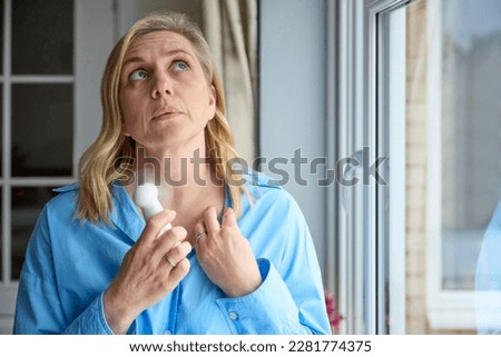 Menopausal Mature Woman Having Hot Flush At Home Cooling Herself With Handheld Electric Fan  Royalty-Free Stock Photo #2281774375