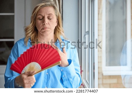 Menopausal Mature Woman Having Hot Flush At Home Cooling Herself With Handheld Paper Fan  Royalty-Free Stock Photo #2281774407