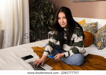 Photo portrait of delighted asian kazachstan girl lady smiling looking at camera in progress buying new items doing purchaase shopping online holding new modern credit card in hand in front of laptop.