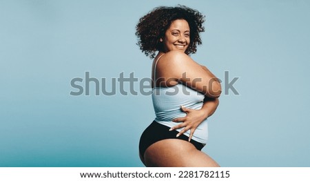Woman looking at the camera as she proudly embraces her body in its natural form. Woman standing confidently in a fitness studio, dressed in sportswear and radiating self-love and acceptance. Royalty-Free Stock Photo #2281782115