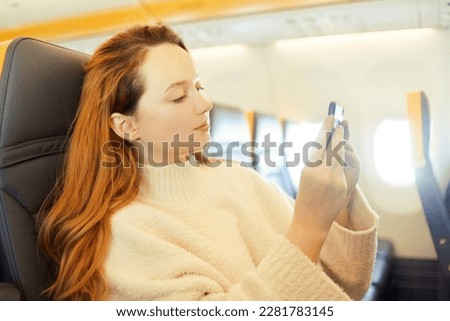A beautiful girl is sitting in an airplane looking at the phone, reading a book or playing games. Air travel, long flight.