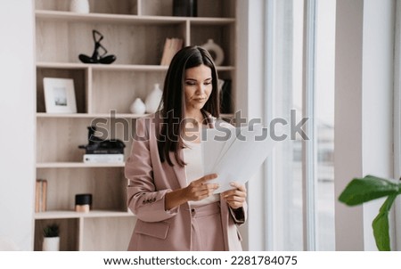 Brunette Asian businesswoman in suit holds sheet of papers looks at documents stands at window against bookshelves. Successful lawyer explores issue at office. Financial bill. Royalty-Free Stock Photo #2281784075