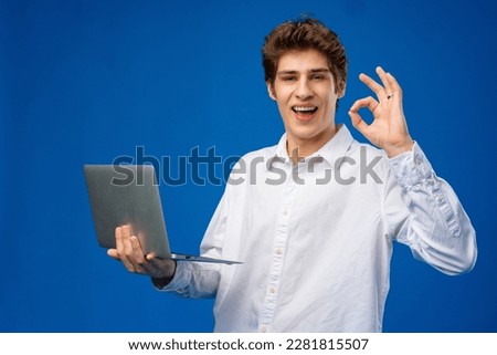 Young man standing in shirt, holding laptop and looking at camera on blue background