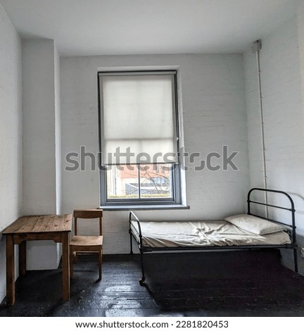 Sparsely decorated room with white walls. Window takes up a large percentage of the back wall. There is a small wooden desk with chair and a bed Royalty-Free Stock Photo #2281820453