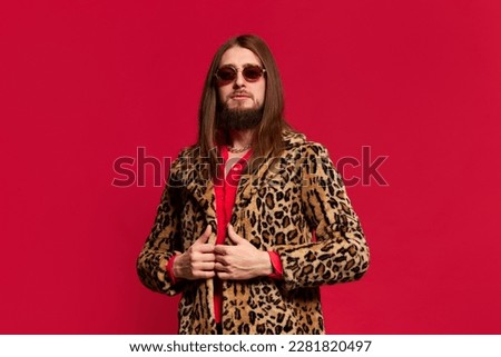 Portrait of young man with long hair posing in stylish fur coat and sunglasses against red studio background. Charismatic look. Concept of emotions, facial expression, lifestyle, unique style Royalty-Free Stock Photo #2281820497