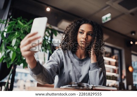 Low angle of smiling young ethnic female touching curly hair with hand and looking at screen of mobile phone while taking selfie and sitting at table with opened planner in blurred cafe