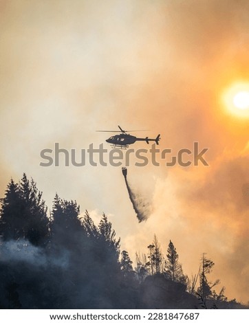 Helicopter attempts to weaken a wildfire. The aircraft flies above the smoke, carrying a bucket of water.