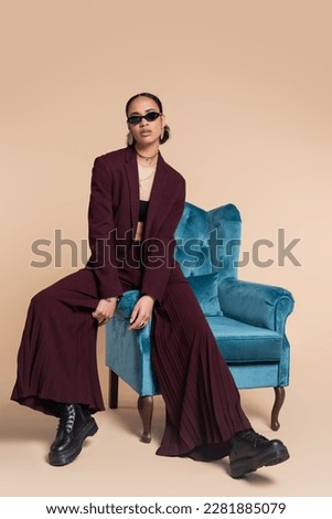 full length of stylish african american woman in burgundy suit and trendy sunglasses sitting on blue velvet armchair on beige