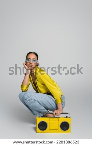 full length of brunette african american woman in blue sunglasses sitting on haunches near yellow boombox on grey Royalty-Free Stock Photo #2281886523