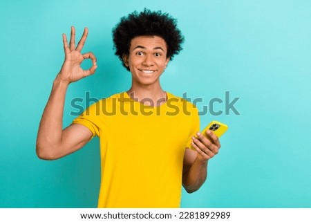Portrait of optimistic satisfied guy wear yellow t-shirt showing okey holding smartphone isolated on vibrant teal color background