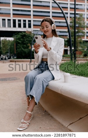Full-length photo of beautiful girl with loose hair wearing white jacket and jeans is listening ,sci, using smartphone and relaxing in city park