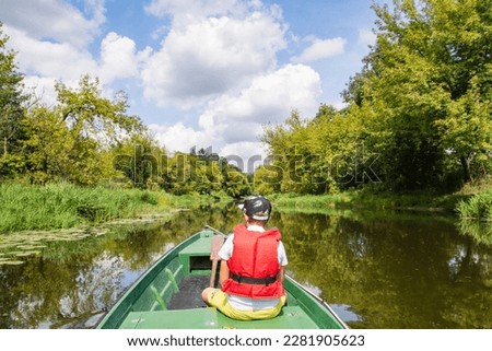 A young boy in a baseball cap and a red life jacket on a green boat is sailing among the trees on a sunny day. Summer.