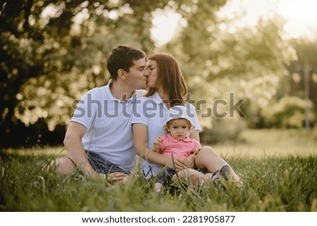 Outdoors, a happy family in nature in summer with a child is sitting on the grass, the summer sun is shining brightly. The couple is kissing
