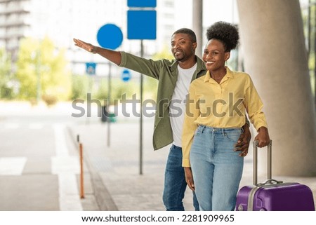 Cheerful African American Spouses With Travel Suitcase Hailing A Taxi Cab With Hand Gesture Standing Outdoors. Tourists Catching Transport Posing Near Modern Airport Outside. Transportation Concept