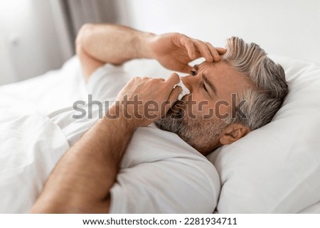 Exhausted sick middle aged grey-haired bearded man wearing pajamas lying in bed with fever and runny nose, touching head and sneezing nose, suffering from cold, flu, coronavirus, closeup Royalty-Free Stock Photo #2281934711