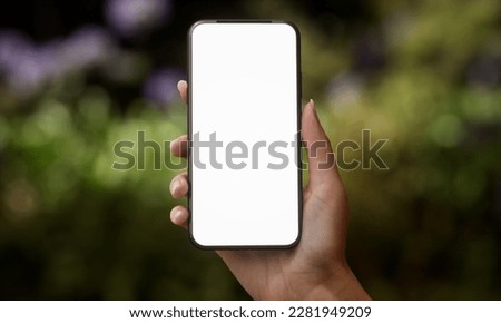 CU Silhouette of Caucasian female holding a generic smart phone in a right hand in vertical orientation, screen facing camera. Garden with green plants in the background Royalty-Free Stock Photo #2281949209