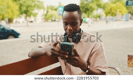 Smiling young african student sits on bench outside of university in headphones, uses phone