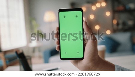 Hand of guys holding a smartphone with green chroma key screen in living room. Template for online store or social media 