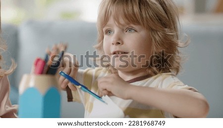 Funny little kid playing while crafting something. Cute boy cutting paper with scissors for children and happily smiling - happy childhood concept 