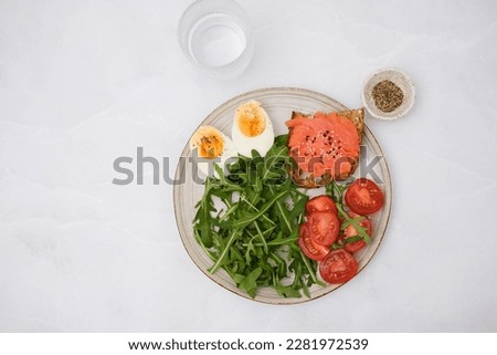 A plate with a salmon sandwiche, arugula, tomatoes, boiled egg with glass of water on marble background. Healthy food. Horizontal frame.