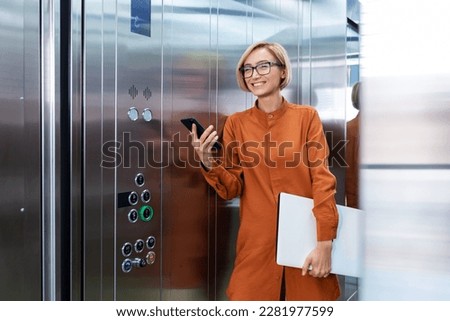 A smiling young business woman is standing in the office center in the elevator, holding a folder with documents and a phone in her hands, going to a conference. meeting. Royalty-Free Stock Photo #2281977599