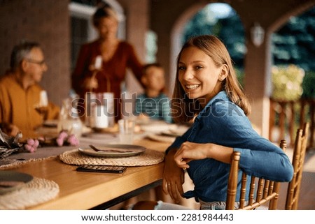 Happy teenage girl at dining table on a patio looking at camera. Her family is in the background.