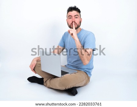 Young caucasian man with laptop sitting over white studio makes silence gesture, keeps finger over lips. Silence and secret concept.