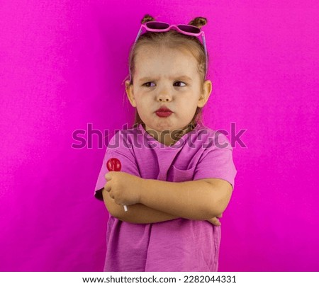 Little child girl pouted with a lollipop in her hands. Child girl cutely pouted her lips and folded her arms on her chest on a pink background. The concept of a funny little girl with a lollipop. Royalty-Free Stock Photo #2282044331