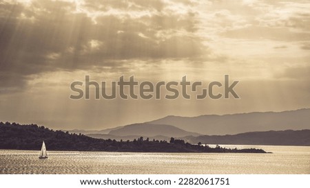 A golden monochrome landscape picture with a sailing boat on a lake, with the mountains in the background and rays of sun  breaking through a cloudy sky