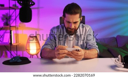 Gamer in neon room. A bearded guy sits at a table and plays a video game on his smartphone. Mobile video games. Rest and relaxation at home.