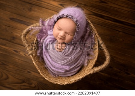 Sleeping newborn baby girl on a background of natural brown wood in a wicker basket. A newborn baby with a lilac-violet cap on his head. Beautiful portrait of a newborn 1-2 weeks 14 days old. Royalty-Free Stock Photo #2282091497