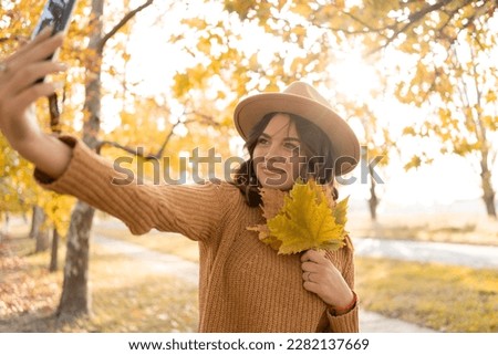 Beautiful young Caucasian woman with fallen leaves taking a selfie with smartphone outdoors in park in autumn. Magnificent brunette girl taking picture of herself while walking. Copy space