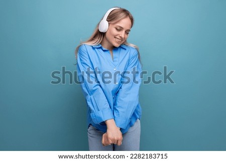 attractive blond girl in a casual shirt listens to songs in big headphones and dances on a blue background