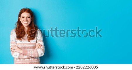 Portrait of happy redhead girl in winter sweater, smiling with arms crossed on chest, standing against blue background.