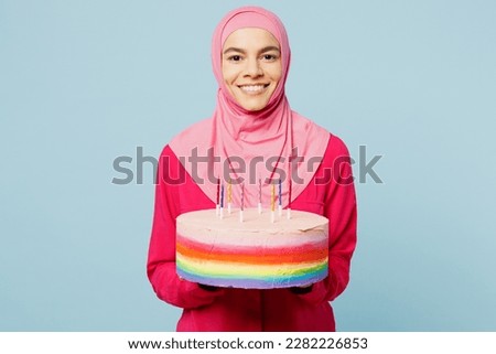 Young happy arabian muslim woman wear pink abaya hijab hold in hand colorful cake with candles isolated on plain pastel light blue cyan background studio portrait. People uae islam religious concept