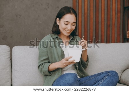 Young fun woman of Asian ethnicity wear casual clothes eat Chinese food cuisine hold takeaway container box sits on grey sofa couch stay home hotel flat rest relax spend free spare time in living room
