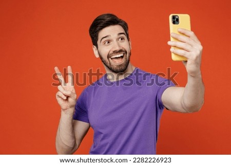 Young man wear casual basic purple t-shirt doing selfie shot on mobile cell phone post photo on social network show v-sign isolated on plain orange background studio portrait. People lifestyle concept
