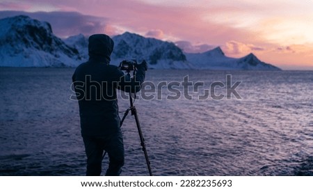 The photographer take a picture of Lofoten islands in winter. A male uses camera and tripod to capture a beautiful landscape picture at twilight in Lofoten. Majestic view of Haukland Beach. 