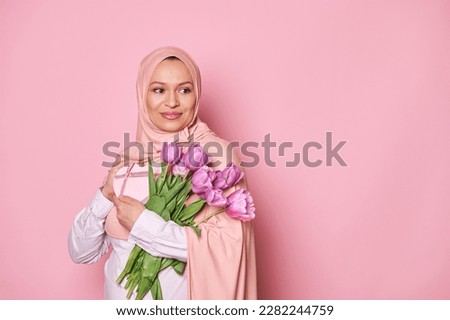 Gorgeous Muslim woman in pink hijab, smiling looking aside, holding a bouquet with purple tulips and a gift box, isolated on pink background. International Women's Day and Mother's day. Free ad space