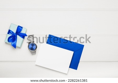 Men's watch with a blue dial, a gift box and a greeting blank with an envelope on a white wooden background. The concept of celebrating father's day, son's day