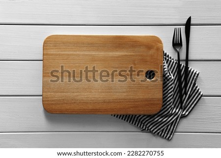 Empty board with cutlery and napkin on white wooden table, flat lay