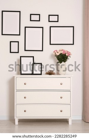 Empty frames hanging on white wall and chest of drawers with flowers in room