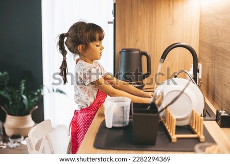 Little girl washing dishes with sponge in kitchen sink at home. Child doing chores concept. Royalty-Free Stock Photo #2282294369