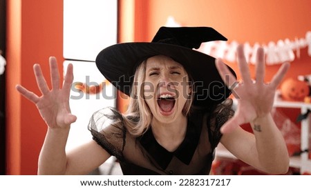 Young blonde woman wearing witch costume standing with scare expression at home