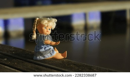 Doll is dressed of blue dress. She has two pigtails. She sits on the wooden bench near the lake. Beautiful girl posing, show direction to the lake by hand. Welcome into deep dive in nature. 