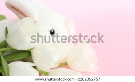 his charming photo features a white toy bunny with adorable eyes sitting next to delicate white tulips. The soft pastel pink background creates a serene and dreamy atmosphere, perfect for Easter or an